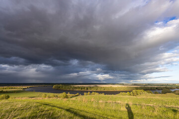 The sky is filled with stormy clouds, casting a moody light over the scene. A person's silhouette is visible on the grassy hill, adding a sense of scale and human presence to the vast natural setting
