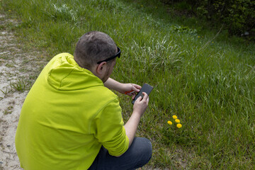  A man in a vibrant yellow shirt and cap is crouched down in a field of green grass and yellow flowers, capturing a photo with his smartphone.