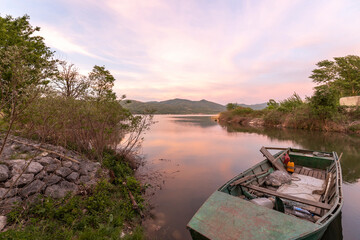 evening scene at a lake with a traditional wooden boat moored at the rocky shore, surrounded by trees and distant mountains under a pink and orange sky