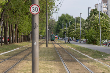  urban park where a green and white tram is in motion on its tracks. The tram is surrounded by lush green trees and grass, with a clear sky overhead.