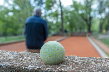 An elderly man is intently focused on a game of boules, with a green boule resting on a stone wall in the foreground. The red court is set in a park with trees, picnic tables, and bicycles