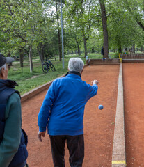 In a serene park setting, two older men engage in a game of boules. One man, dressed in a blue jacket and beige vest, is in the midst of a focused throw, while the other, wearing observes.