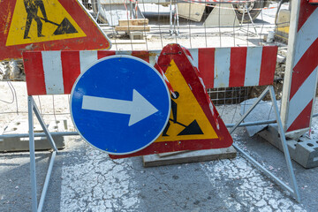 A construction site with a worker symbol and directional arrow sign indicating set against a backdrop of red and white striped barriers, symbolizing caution and a temporary change in road direction