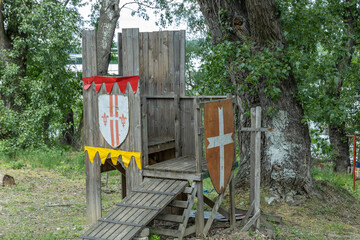 A rustic wooden structure resembling a medieval fortification or play area, featuring a throne-like seat, a shield with a cross, and a red banner. 