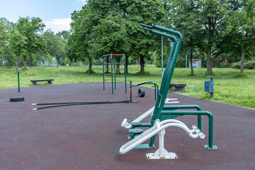 An outdoor fitness park featuring a variety of exercise equipment such as pull-up bars, leg press machine, and push-up bars, set on a rubber flooring surface with a backdrop of trees.