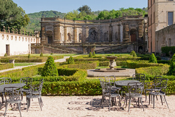 A beautiful summer park with figured trees and paths in the courtyard of an ancient villa, fenced with an ancient stone wall with sculptures, there is figured garden furniture made of metal