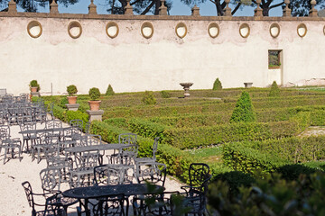 A beautiful summer park with figured trees and paths in the courtyard of an ancient villa, fenced with an ancient stone wall with sculptures, there is figured garden furniture made of metal