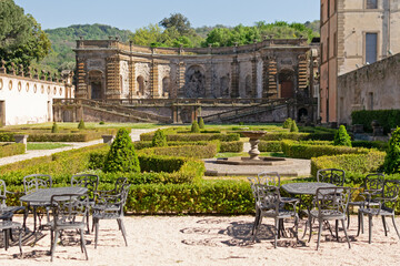 A beautiful summer park with figured trees and paths in the courtyard of an ancient villa, fenced with an ancient stone wall with sculptures, there is figured garden furniture made of metal