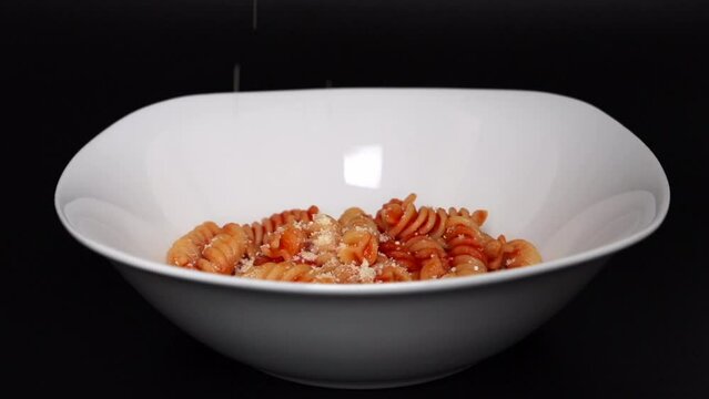 Side view of sprinkling parmesan cheese into fusilli pasta with tomato sauce in white bowl on the black background 