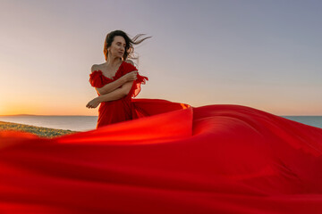 woman red dress standing grassy hillside. The sun is setting in the background, casting a warm glow...