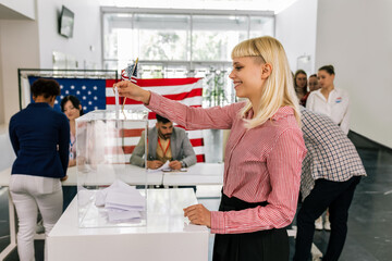 Young blonde woman voter placing ballot in ballot box polling place in America.