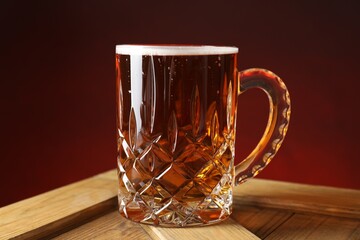 Mug with fresh beer on wooden crate against burgundy background, closeup