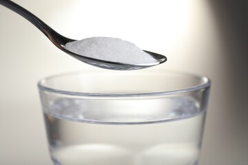 Spoon with baking soda over glass of water on light background, closeup