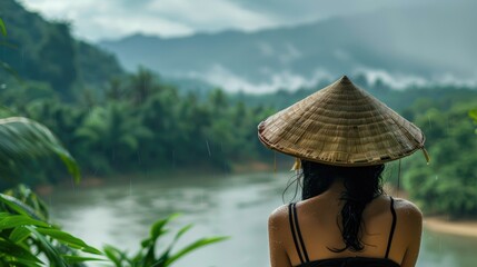 A woman sporting a traditional bamboo hat gazes out at the river and lush rainforest from her vantage point capturing the essence of the rainy season on a tropical island This picturesque s