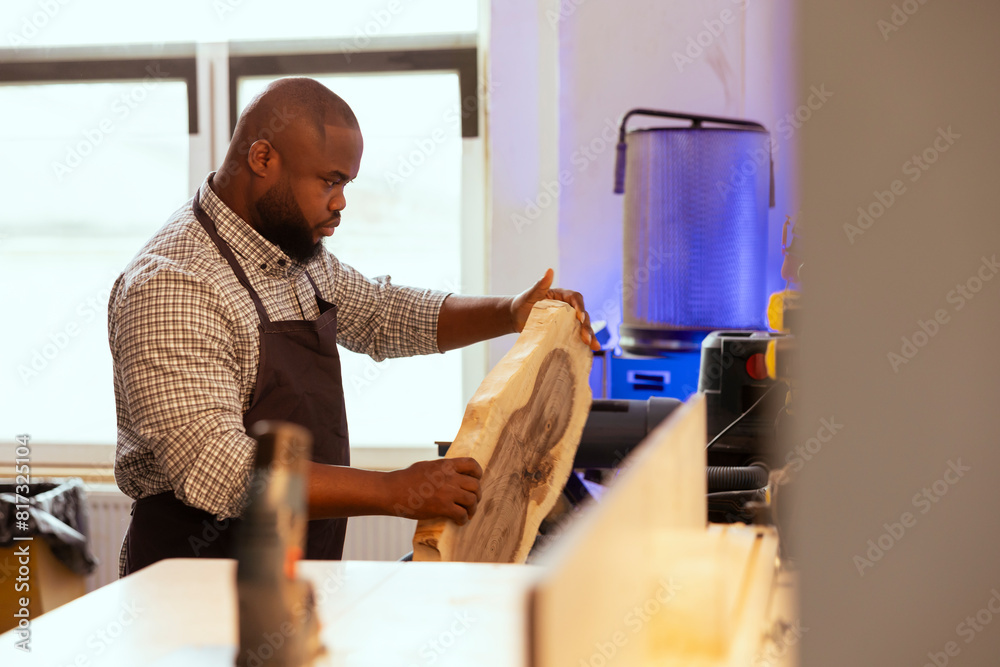 Wall mural African american carpenter in woodworking shop inspecting wood before assembling furniture. BIPOC cabinetmaker in joinery studio evaluating lumber block, checking for damages