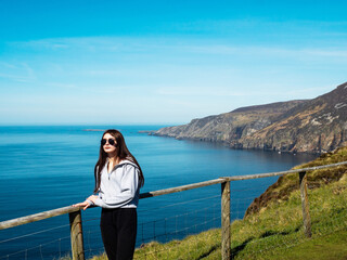 Teenager girl on a trip to Slieve League Cliff, Ireland. Stunning nature scenery with cliff, ocean and clean blue sky. Travel and tourism concept. Model posing in nature scene. Sunny day.
