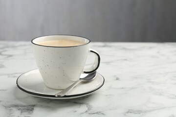 Tasty cappuccino in cup, spoon and saucer on white marble table, closeup. Space for text