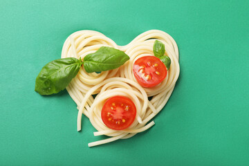 Heart made of tasty spaghetti, tomato and basil on green background, top view