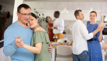 Happy middle-aged men and women doing slow dance in pairs during cozy get-together with friends