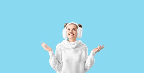 Happy young woman in warm sweater and with fluffy headphones on light blue background