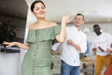 Cheerful young Asian woman in green off-shoulder dress enjoying lighthearted active dancing with friends of different nationalities at casual relaxed home gathering