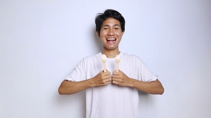 excited young asian man in white shirt gestures holding spoon and fork made of bamboo