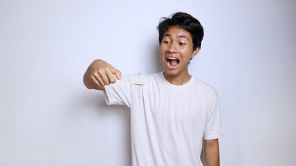 Excited young Asian man in white shirt gestures while eating holding a spoon made of bamboo