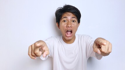 young asian man in white shirt with funny expression shocked gesture pointing forward, looking at...