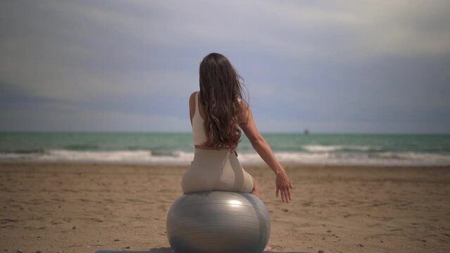 A Latina woman sits on an exercise ball, stretching and relaxing on a sandy beach with the ocean in the background.