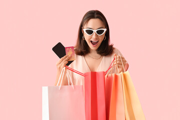 Young woman with credit card, mobile phone and shopping bags on pink background