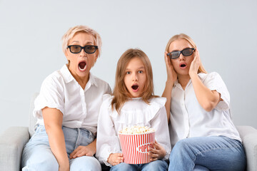 Shocked little girl with her mom and grandmother watching movie on sofa against light background