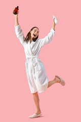 Happy young woman in bathrobe with bottles of shower gel on pink background