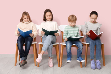 Little children reading books while sitting on chairs near pink wall