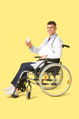 Young male doctor in wheelchair with laptop and pills on yellow background