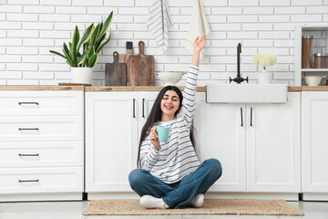 Cheerful young woman with cup of tea sitting on rug in light kitchen
