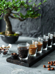 Glass cups with different shades of coffee are stacked on a black wooden board, with chocolate-covered hazelnuts and a modern bonsai tree nearby.Minimal creative drink and food concept.