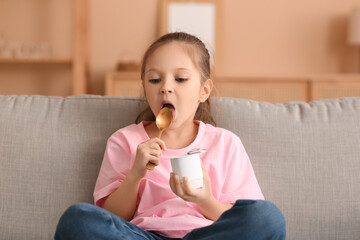 Cute little girl sitting on sofa and eating yogurt at home