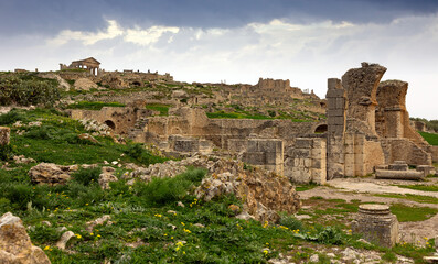 Obraz premium Scenic view of Licinius Sura Baths, well-preserved Roman ruins in Dougga, surrounded by spring greenery in Tunisia against cloudy backdrop