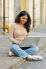 Indian woman working online distant job with laptop sitting on city street browsing website chatting outdoors during break. Girl looking at notebook screen send messages watching movies. Vertical