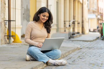 Indian woman freelancer working online distant job with laptop sitting on city street browsing website chatting outdoors during break. Girl looking at notebook screen send messages watching movies.