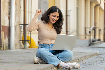 Overjoyed happy young Indian woman working on laptop scream in delight raise hands in triumph winner gesture celebrate success game money win outdoors. Happy girl sitting on downtown street in city