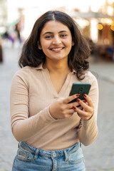 Happy smiling Indian woman using smartphone typing text messages browsing internet, finishing work, looking at camera outdoors. Hispanic girl walking on urban city street. Town lifestyles. Vertical