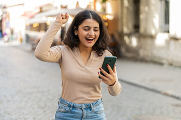 Indian woman use mobile smartphone celebrating win good message e-mail news, lottery jackpot victory, giveaway online outdoors. Hispanic girl walking on urban city street. Town lifestyles