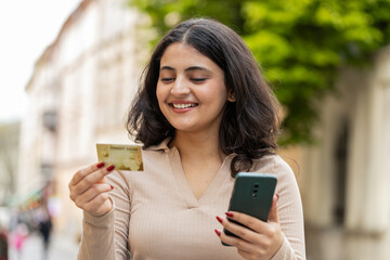 Indian young woman using credit bank card smartphone while transferring money, purchases online shopping, order food delivery, booking hotel room outdoors. Hispanic girl walking on urban city street