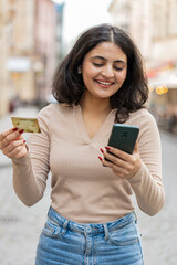 Indian young woman using credit bank card smartphone while transferring money, purchases online shopping, order food delivery, booking hotel room outdoors. Girl walking on urban city street. Vertical