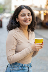 Smiling happy Indian young woman enjoying morning coffee hot drink outdoors. Relaxing taking a break. Girl drinking coffee to go walking passes the urban city street. Town lifestyles outside. Vertical