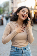 Phone call good news gossip. Happy surprised amazed Indian woman in pleasant conversation on smartphone enjoying talking with friends outdoors. Hispanic girl walking on urban city street. Vertical