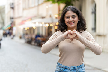 I love you. Indian young woman makes symbol of love, showing heart sign to camera, express romantic feelings, express sincere positive feelings. Charity, gratitude, donation. Girl on urban city street