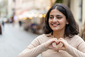 I love you. Indian young woman makes symbol of love, showing heart sign to camera, express romantic feelings, express sincere positive feelings. Charity, gratitude, donation. Girl on urban city street