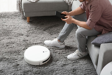 Man with mobile phone and modern robot vacuum cleaner sitting on sofa at home